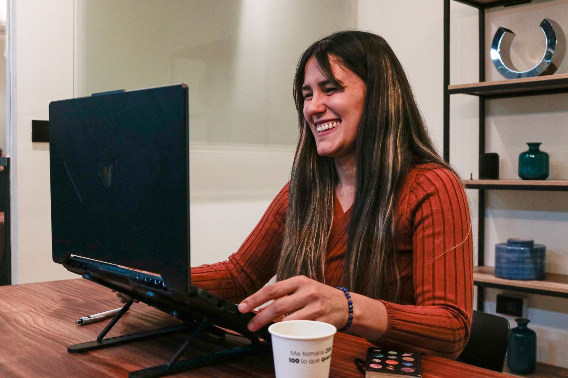 Smiling woman in red sweater enjoying a video call at home office setup with laptop and coffee.