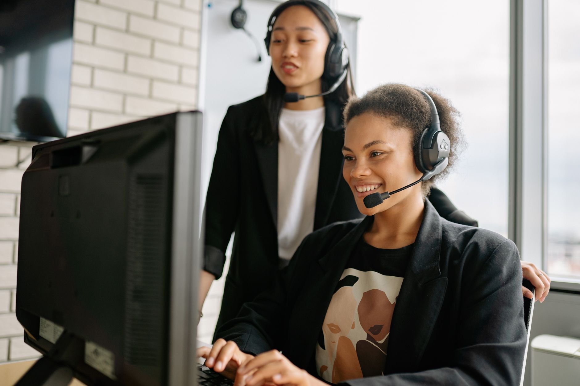 Two women with headsets engaged in customer service at a computer in a bright office.